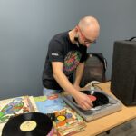 DJ przygotowuje gramofonu do odtwarzania, na stole leżą różne płyty winylowe
