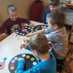 Cztery chłocy z uśmiechami robią zegary, kleją kolorowe kółeczka z numerami, wskazówki.