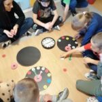 Na podłodze dzieci robią zegary, kleją kolorowe kółeczka z numerami, wskazówki. W środku leży prawdziwy zegar. Jeden już jest gotowy i pokazuje godzinę drugą.