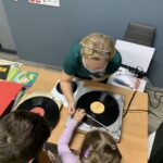Dziewczynka z pomocą bibliotekarki ustawia głowicy gramofonu.