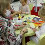 Widok na stół i ręce dzieci robią marakasy z grubej tektury i różnych materiałów. Pomaga jej babcia.