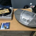 Na stole leżą: gramfon odtwarzający pocztówkę dźwiękową, odtwarzacz płyt CD i etui na płytę CD z muzyką Marka Grechuty